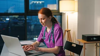 A woman in scrubs focused on her laptop, engrossed in her work.